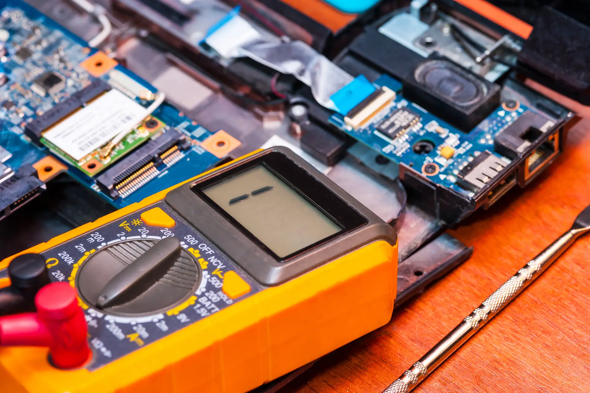Gelbes Multimeter mit neutraler Anzeige neben Laptopplatine und Schraubendreher